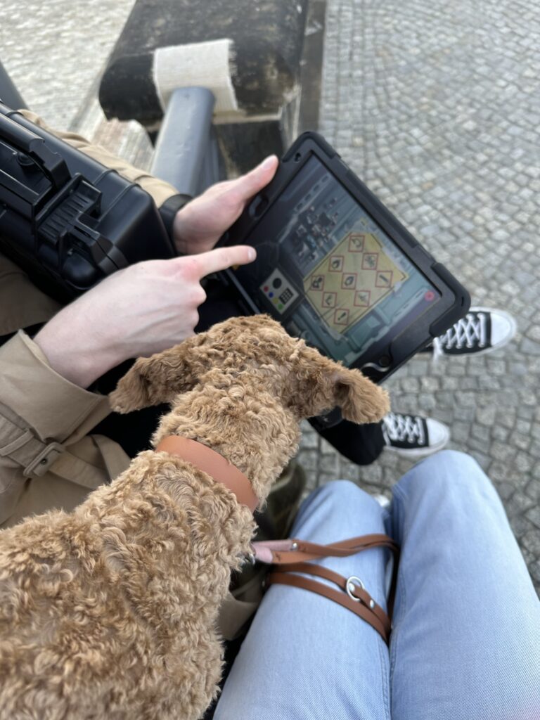 Ein Pudel guckt auf ein Tablet. Auf dem Tablet läuft ein Escape Game und ein Finger von einem Mann tippt darauf.